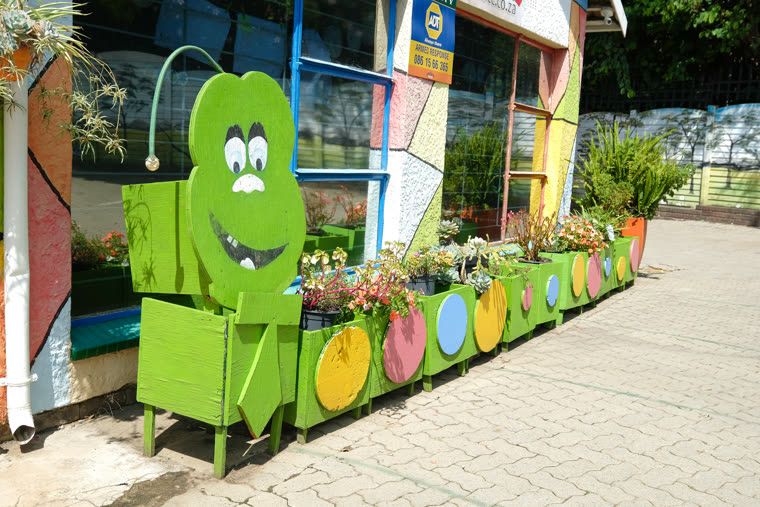 A paved path along the classrooms, with a green caterpillar planter and painted stepping-stone shapes on the wall